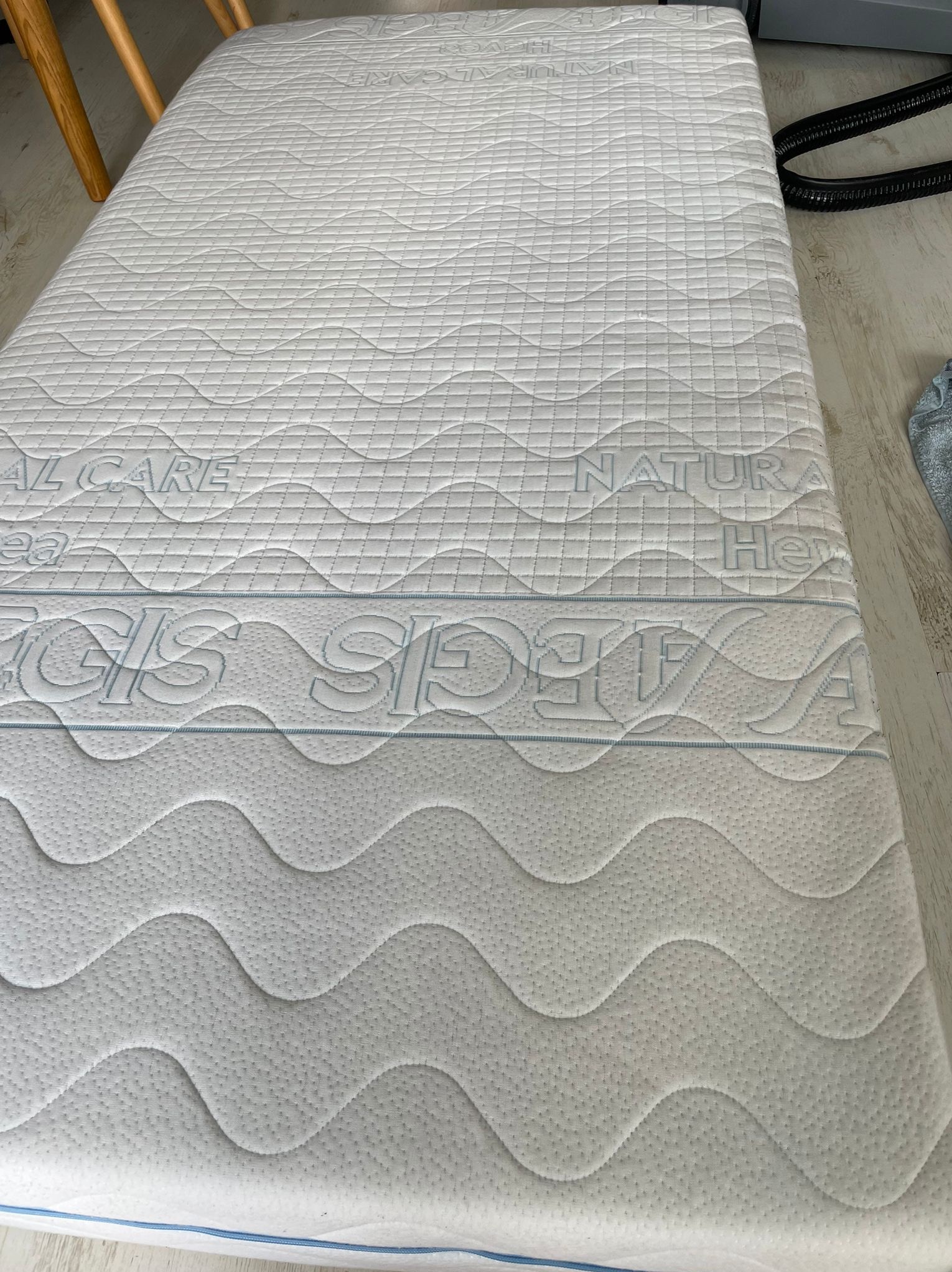 Biały materac z wzorem fal i napisem 'NATURAL CARE' oraz 'REGLIS SLEEP' w górnej części, leżący na podłodze, ukazujący strukturę i detale materiału.