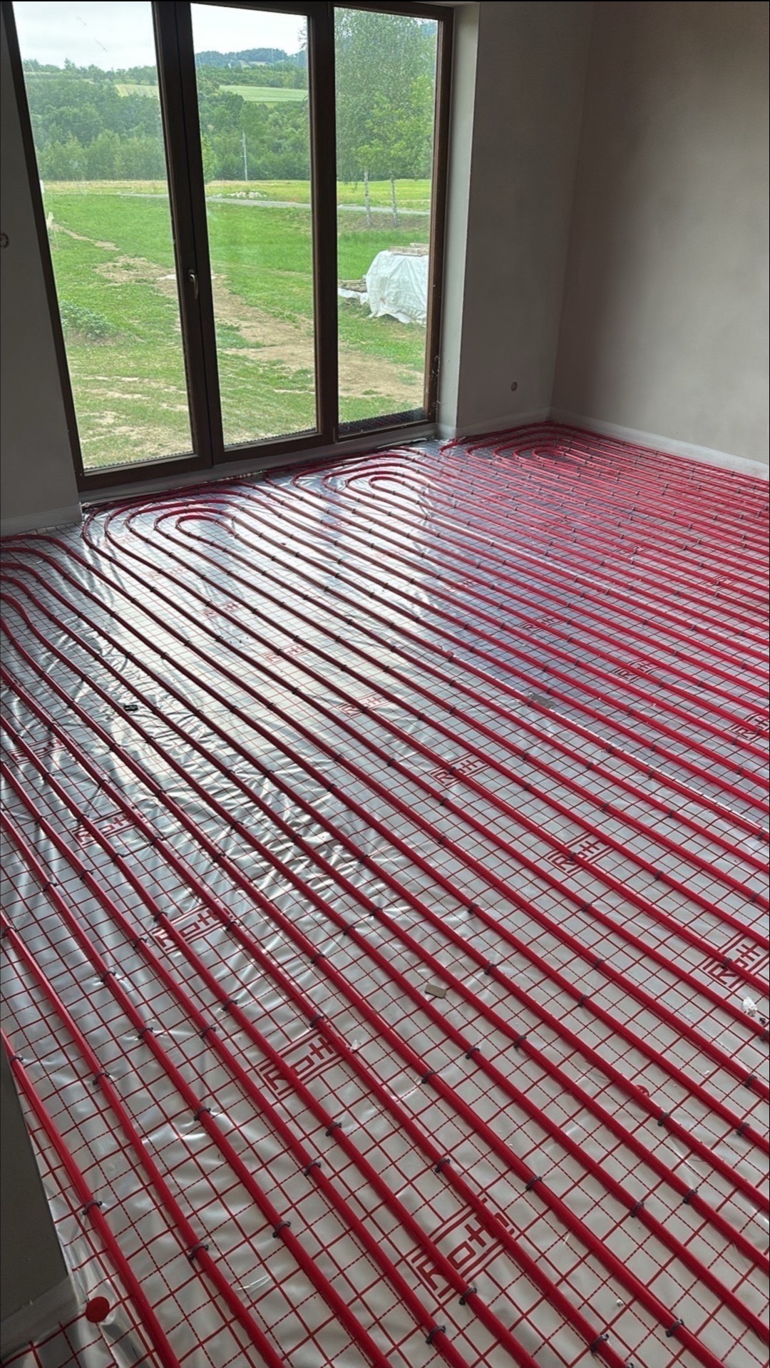 Czerwone rury ogrzewania podłogowego ułożone na folii, widok z góry, w tle okno z widokiem na zieleń, przygotowane do zalania posadzką.