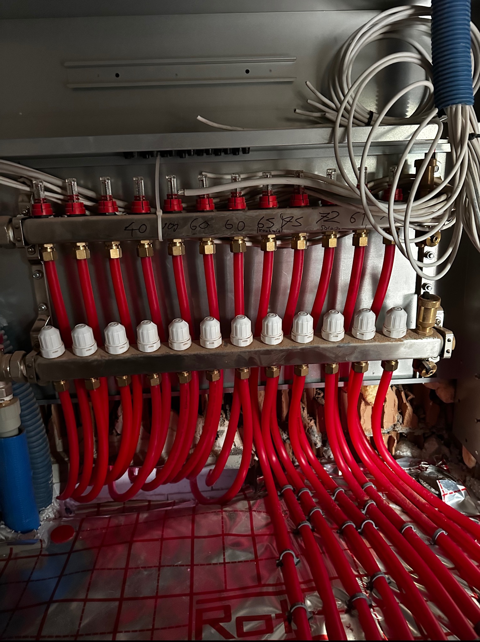 Rozdzielacz ogrzewania podłogowego z czerwonymi rurami na folii z nadrukowaną siatką, widoczne numery obwodów, całość w szarej szafce instalacyjnej z przewodami.