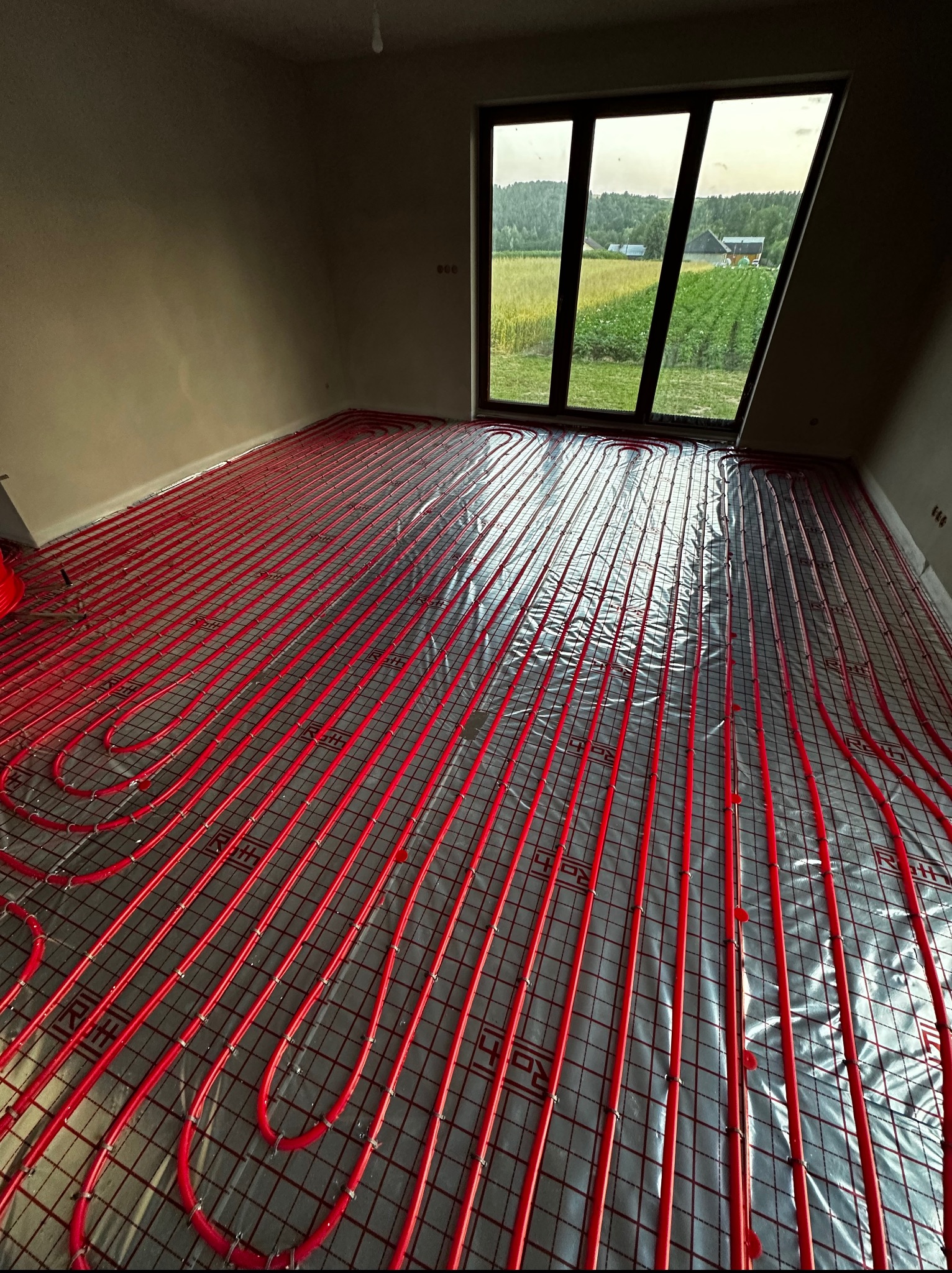 Czerwone rury ogrzewania podłogowego ułożone na folii w jasnym pokoju z dużym oknem, przez które widać zielony krajobraz. Widoczne elementy montażowe.