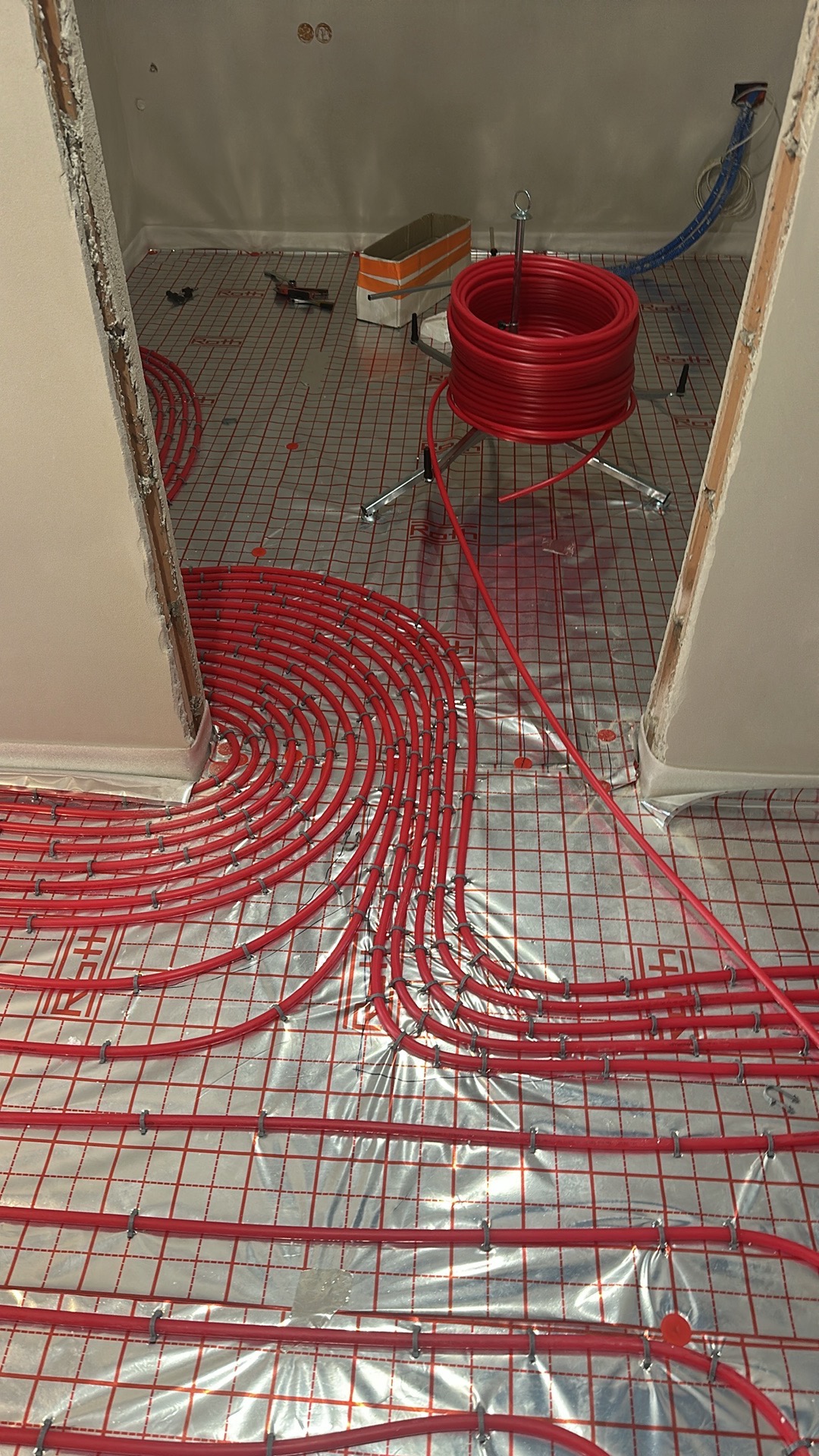 Czerwone rury ogrzewania podłogowego, rozłożone na folii z siatką, w trakcie montażu. Widoczna rolka rur na stojaku i fragment ściany w surowym stanie.