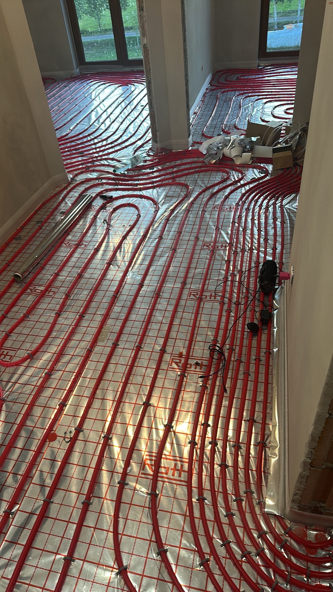 Czerwone rury ogrzewania podłogowego, ułożone na folii z siatką, w surowym wnętrzu. Widoczne okno i przygotowane materiały montażowe.