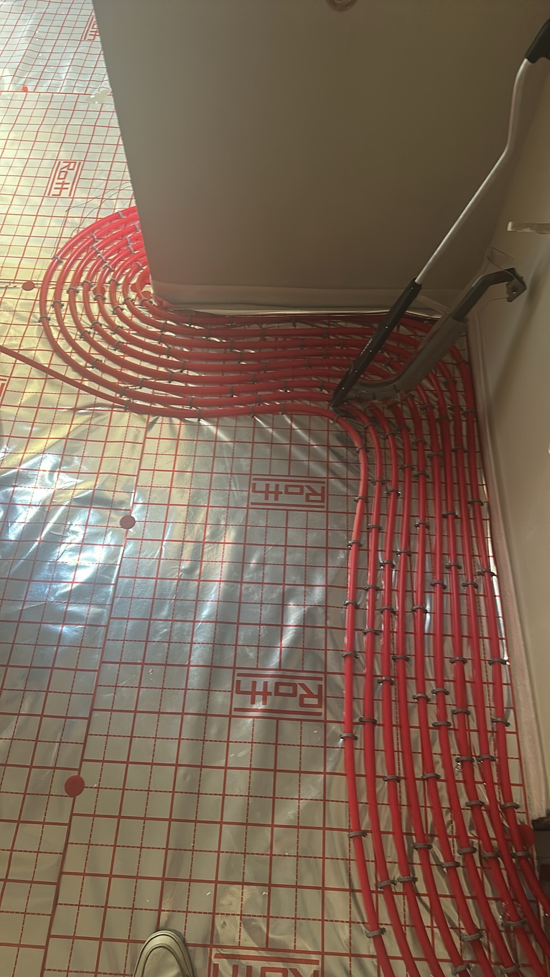 Czerwone rury ogrzewania podłogowego Roth, równomiernie rozłożone na izolacyjnej macie z siatką, przygotowane do zalania. Widoczne mocowania i fragment podłogi.