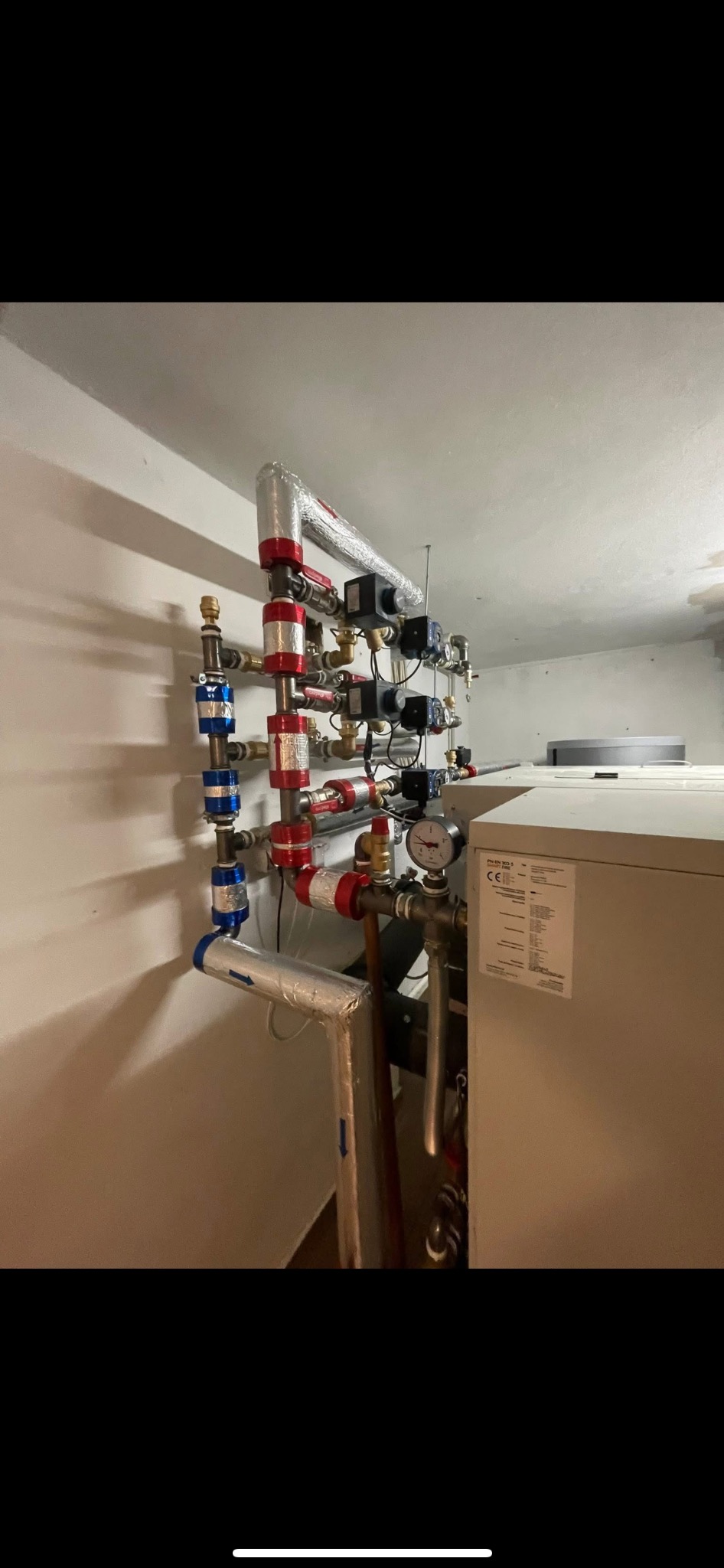 Złożona instalacja grzewcza z rurami w kolorze czerwonym i niebieskim, z zaworami i manometrem, zamontowana na ścianie w piwnicy obok pieca.