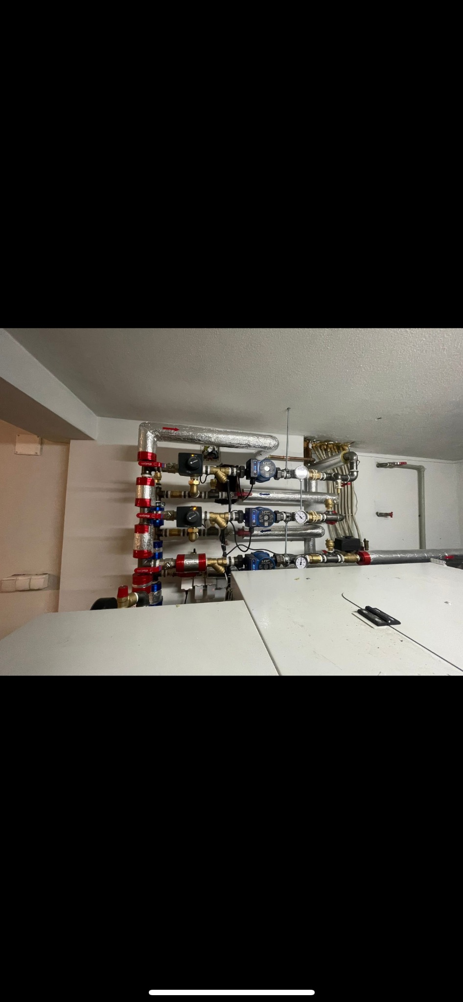 Instalacja grzewcza z licznymi rurami, zaworami i pompami w piwnicy. Elementy w kolorach czerwonym, niebieskim i srebrnym. Widoczne manometry i izolacje termiczne.