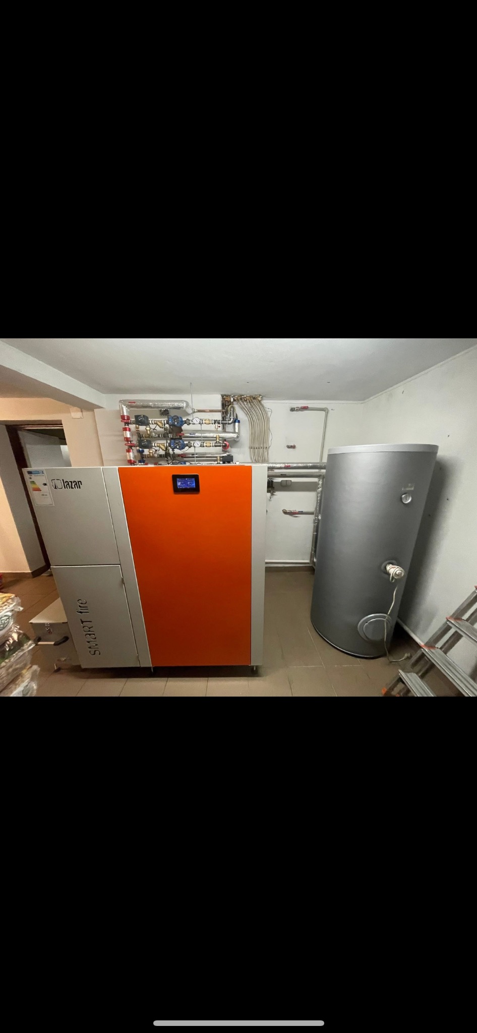 Nowoczesny piec Lazar Smart Fire w kotłowni, z szarym zbiornikiem buforowym i drabinką. Widoczne rury instalacji grzewczej na ścianie.