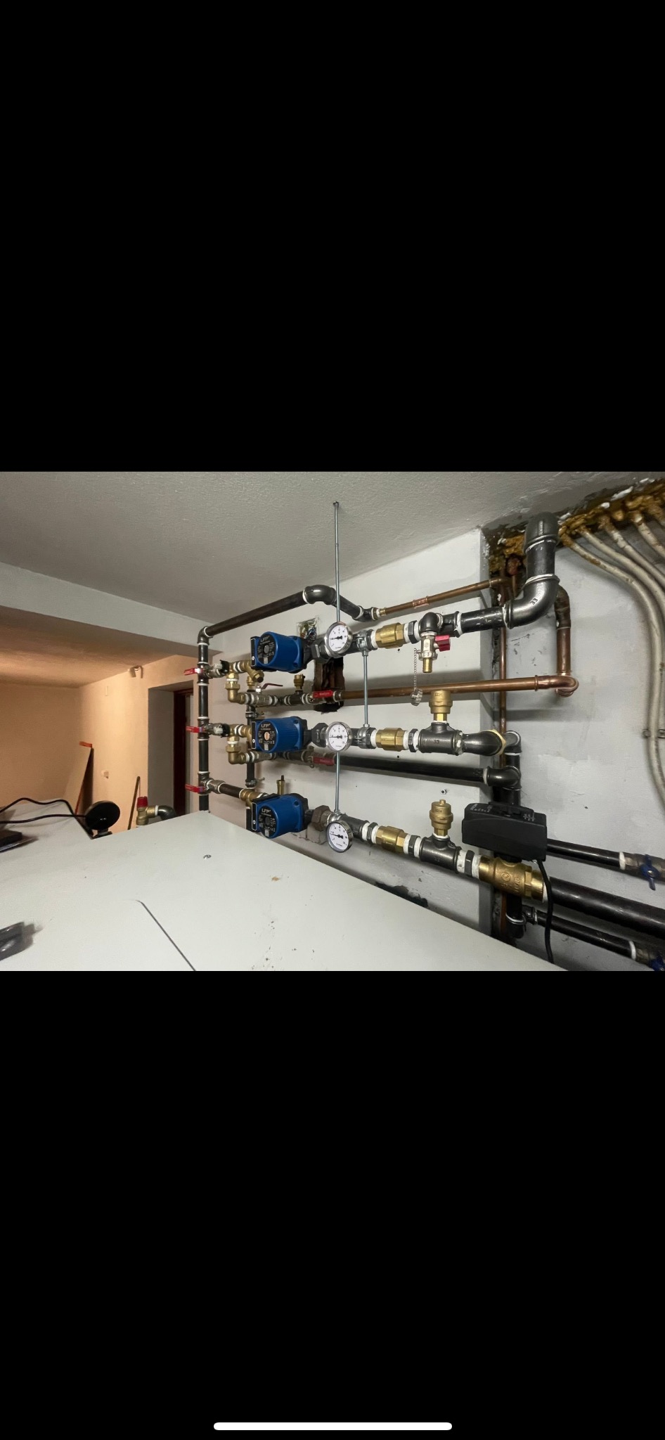 Rozbudowana instalacja grzewcza z pompami obiegowymi, manometrami i zaworami, zamontowana na ścianie w piwnicy. Widoczne rury miedziane i stalowe.