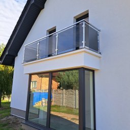 AP-STAL - Nowoczesny balkon z szklaną balustradą i stalowymi słupkami, widok z dołu, na tle białej elewacji i błękitnego nieba.