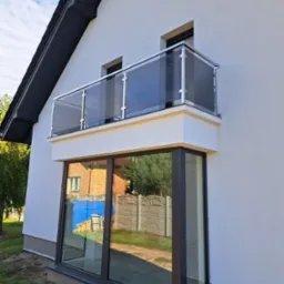 Nowoczesny balkon z szklaną balustradą i stalowymi słupkami, widok z dołu, na tle białej elewacji i błękitnego nieba.