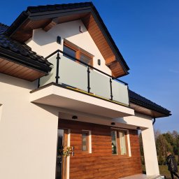 AP-STAL - Dom z balkonem zabezpieczonym szkłem mlecznym, czarne obramowanie. Elewacja w bieli i drewnie. W tle błękitne niebo i zieleń drzew. Ujęcie z dołu.