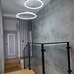 AP-STAL - Nowoczesna balustrada szklana ze szczeblami i czarnymi elementami mocującymi, oświetlona okrągłymi lampami LED na schodach z drewnianymi stopniami i szarą ścianą.