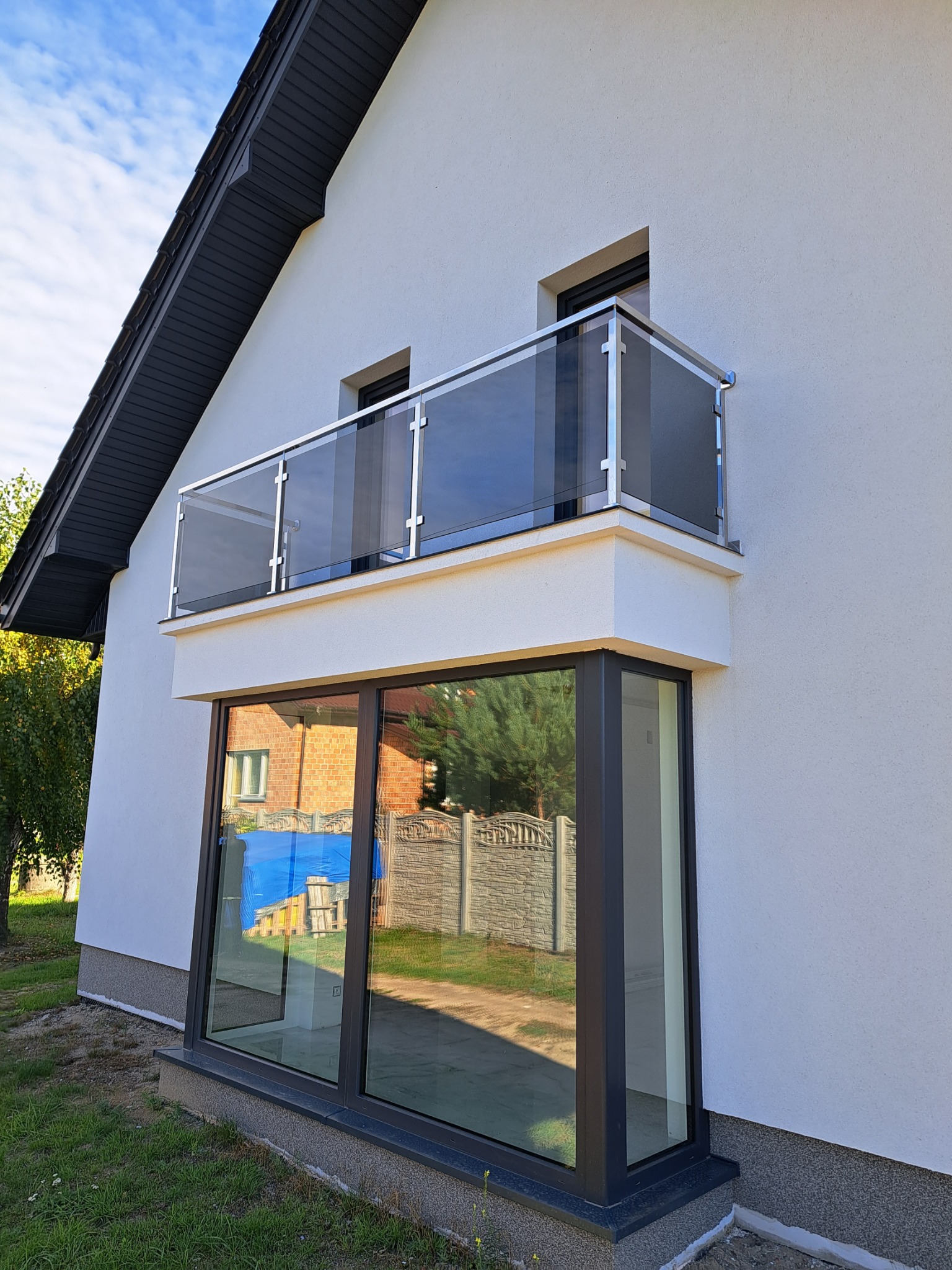 Nowoczesny balkon z szklaną balustradą i stalowymi słupkami, widok z dołu, na tle białej elewacji i błękitnego nieba.