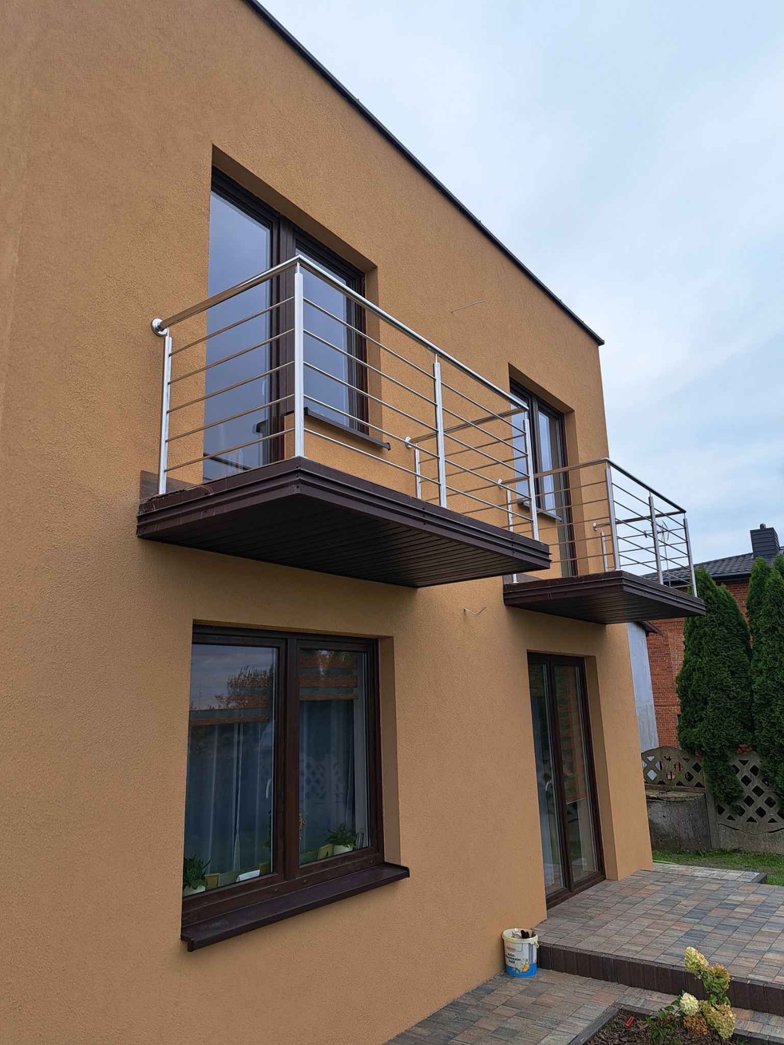 Dwa balkony z metalową konstrukcją na elewacji domu o piaskowym odcieniu. Uwagę przykuwa nowoczesny design i proste linie balkonów.