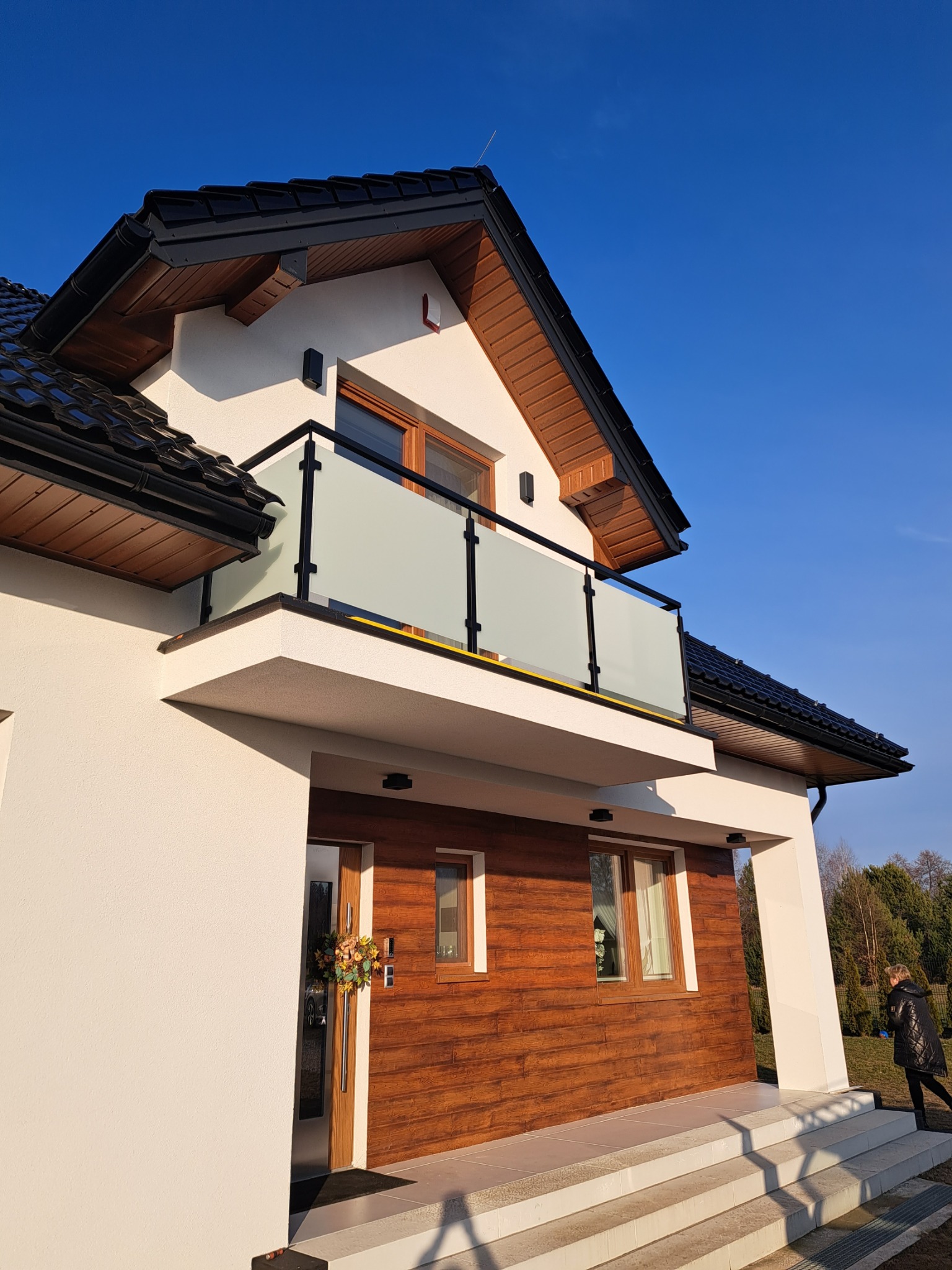 Dom z balkonem zabezpieczonym szkłem mlecznym, czarne obramowanie. Elewacja w bieli i drewnie. W tle błękitne niebo i zieleń drzew. Ujęcie z dołu.