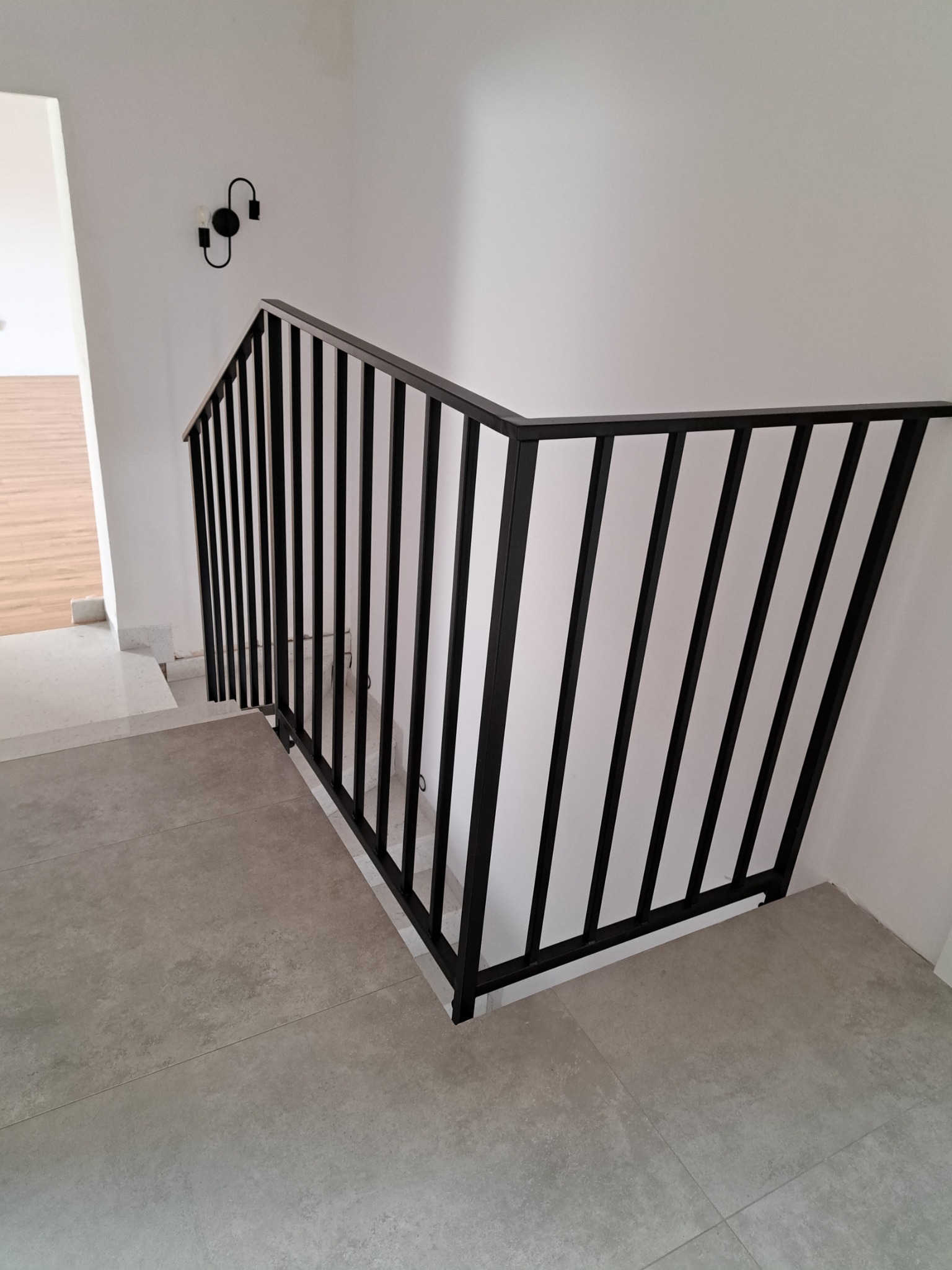 Nowoczesna, czarna poręcz schodowa z pionowymi szczeblami, zamontowana na jasnych schodach w minimalistycznym wnętrzu. Widoczny fragment lampy ściennej.