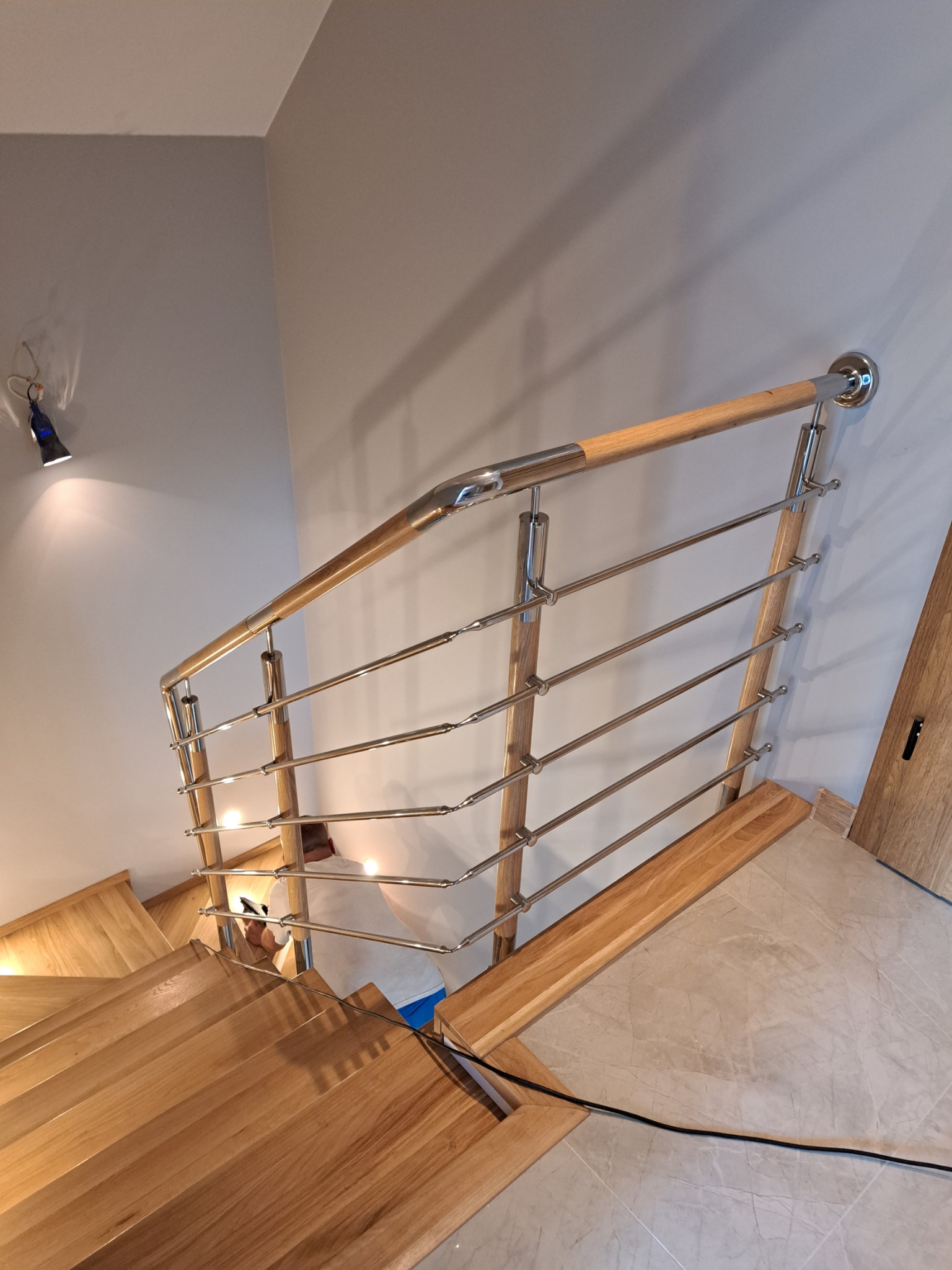 Poręcz schodowa z drewna i stali nierdzewnej, mocowana do ściany, widok z dołu na drewniane schody i fragment podłogi z szarych płytek.