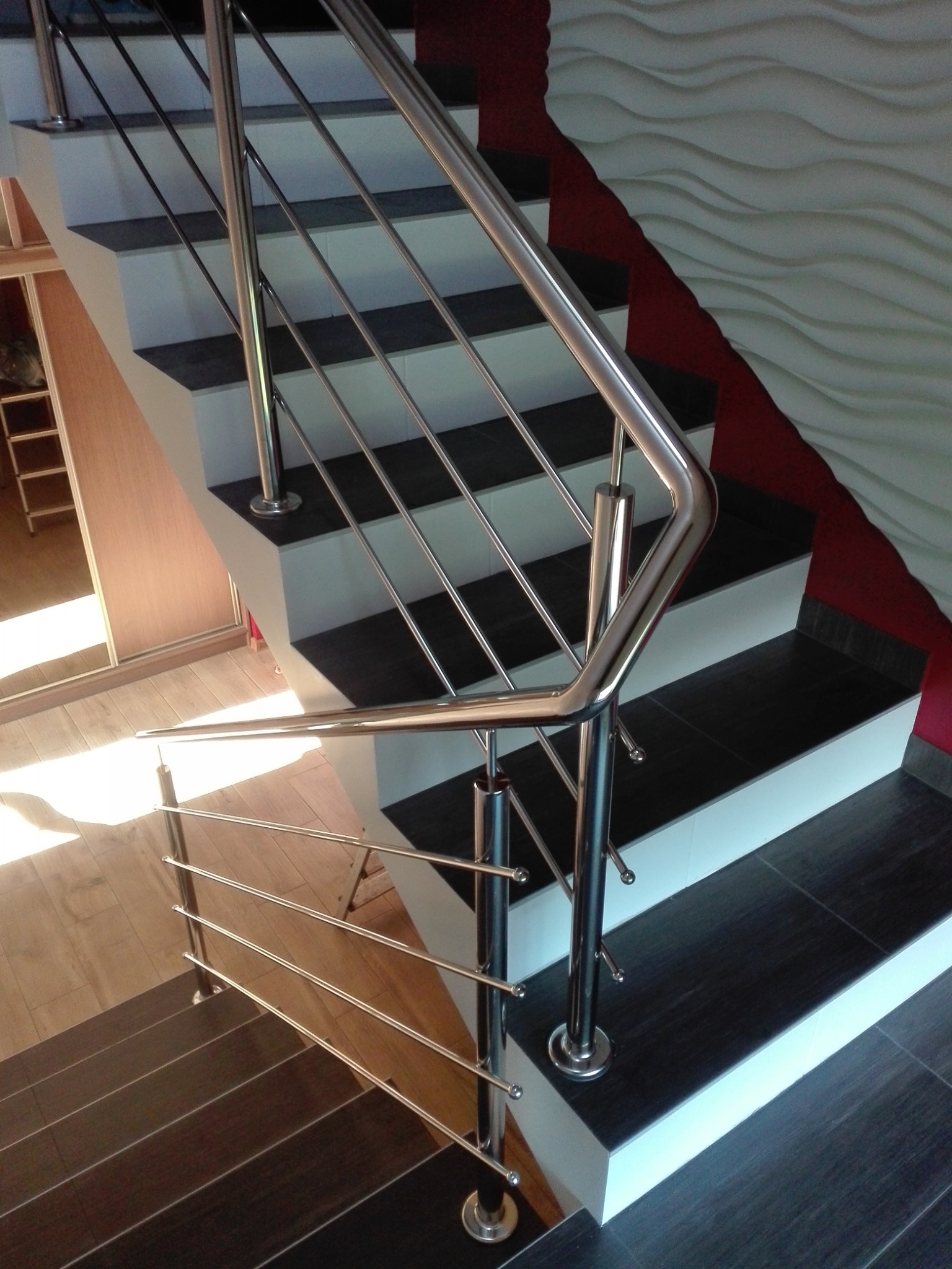 Nowoczesne schody z połyskującą, metalową poręczą z poziomymi prętami. Ciemne stopnie kontrastują z białymi podstopnicami i dekoracyjną ścianą.