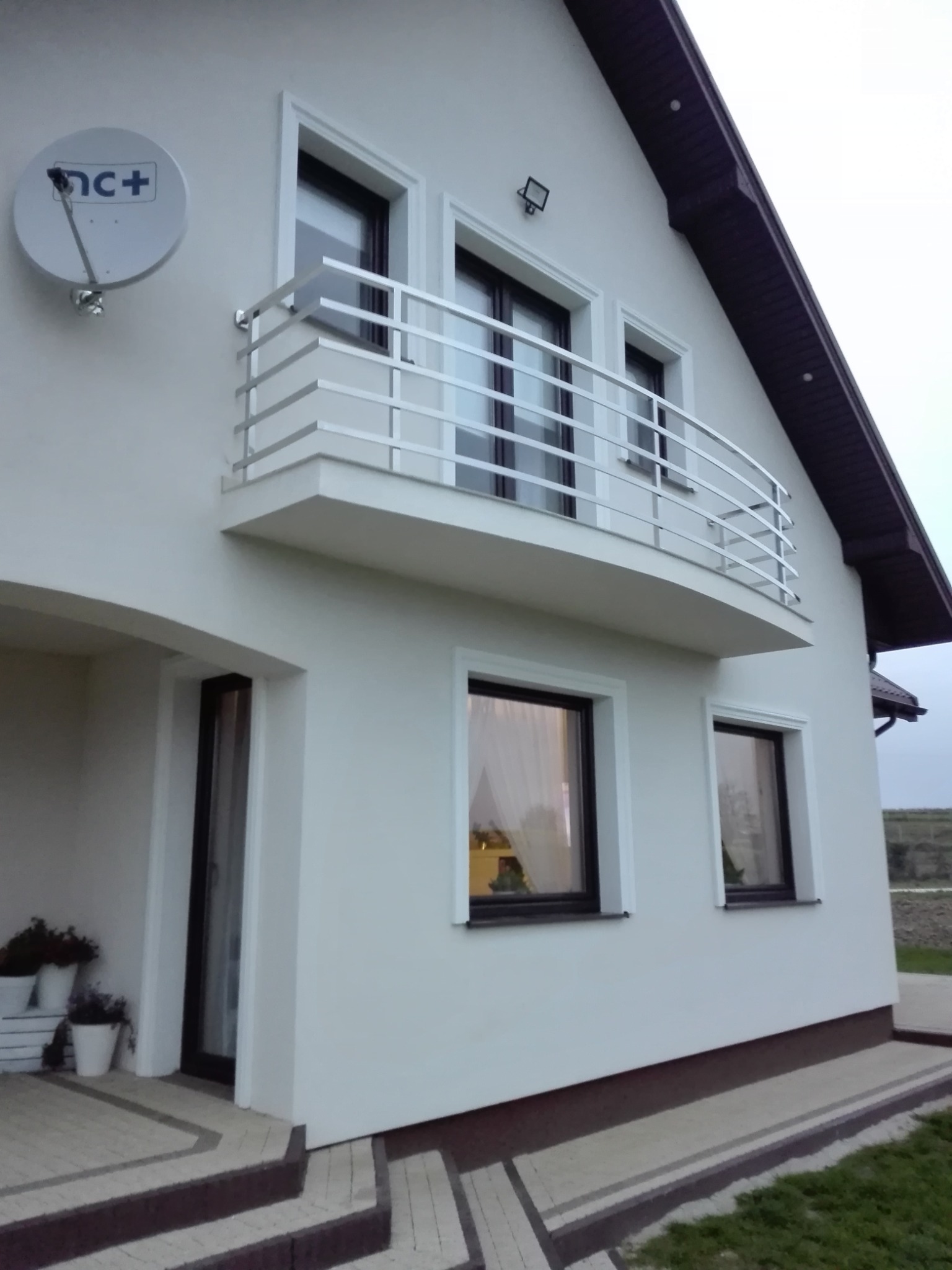 Biały dom z balkonem i nowoczesną, metalową balustradą z poziomych prętów. Widoczna antena satelitarna i okna z ciemnymi ramami.