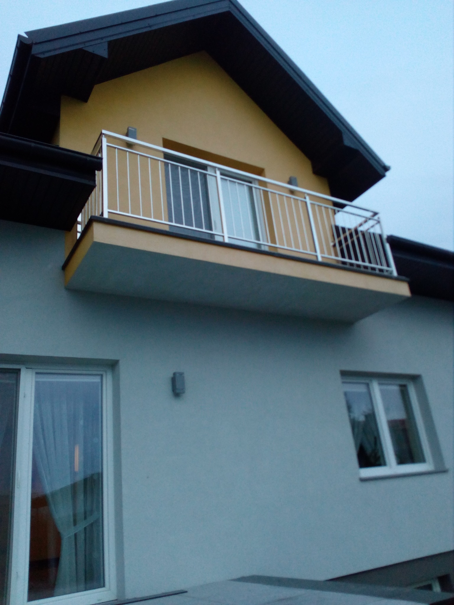Dom z balkonem zabezpieczonym nowoczesną, jasną balustradą z cienkich, pionowych prętów. Widok z dołu na fragment elewacji i wystający balkon.