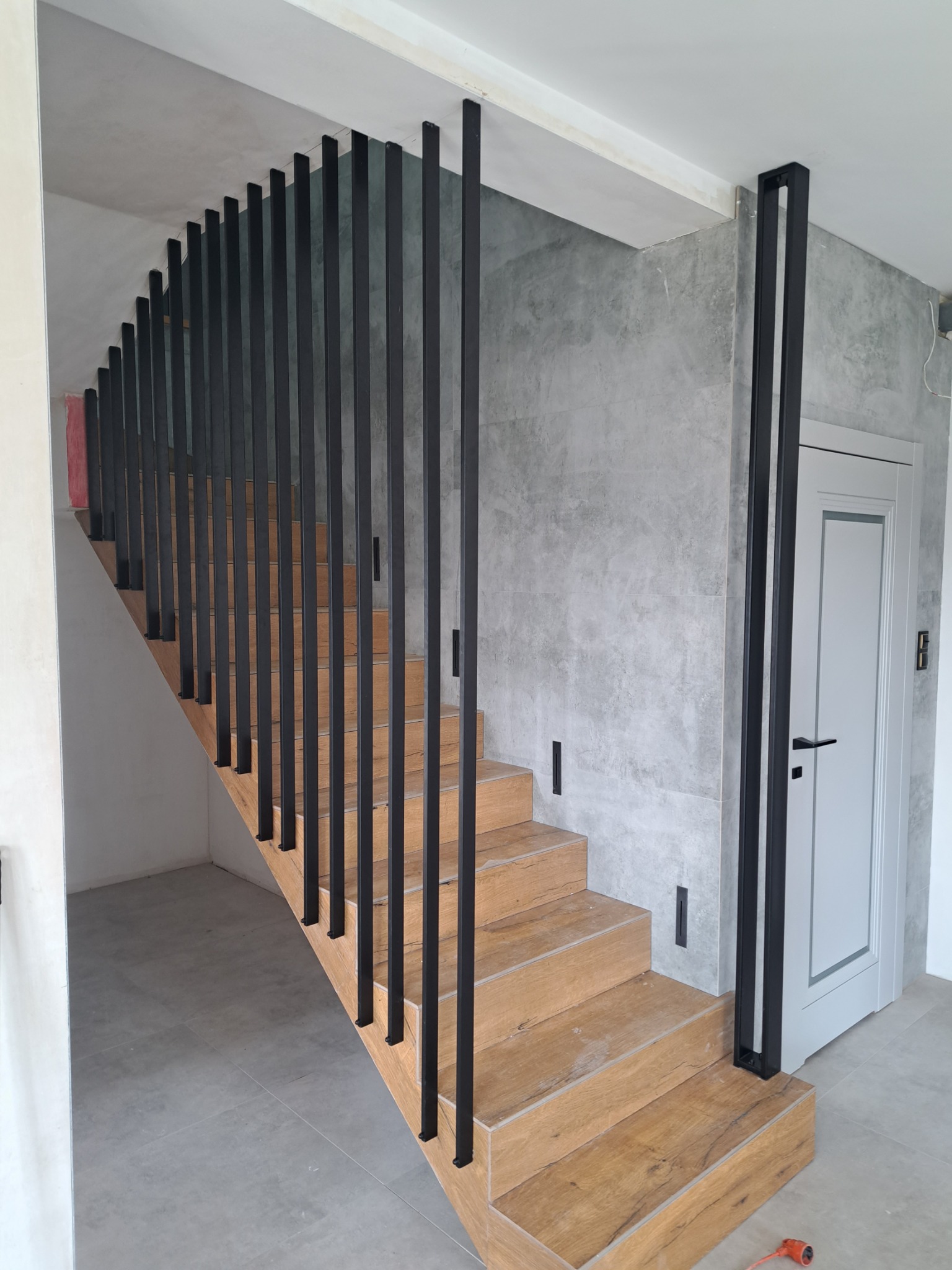 Nowoczesna balustrada z czarnych, pionowych słupków na drewnianych schodach w szaro-białym wnętrzu. Widoczne minimalistyczne detale i oświetlenie.