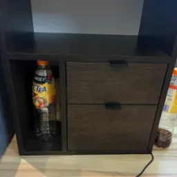 Ciemna szafka nocna z dwiema szufladami i otwartą półką, na której stoi butelka Ice Tea. Na blacie szafki widoczny różowy pojemnik.