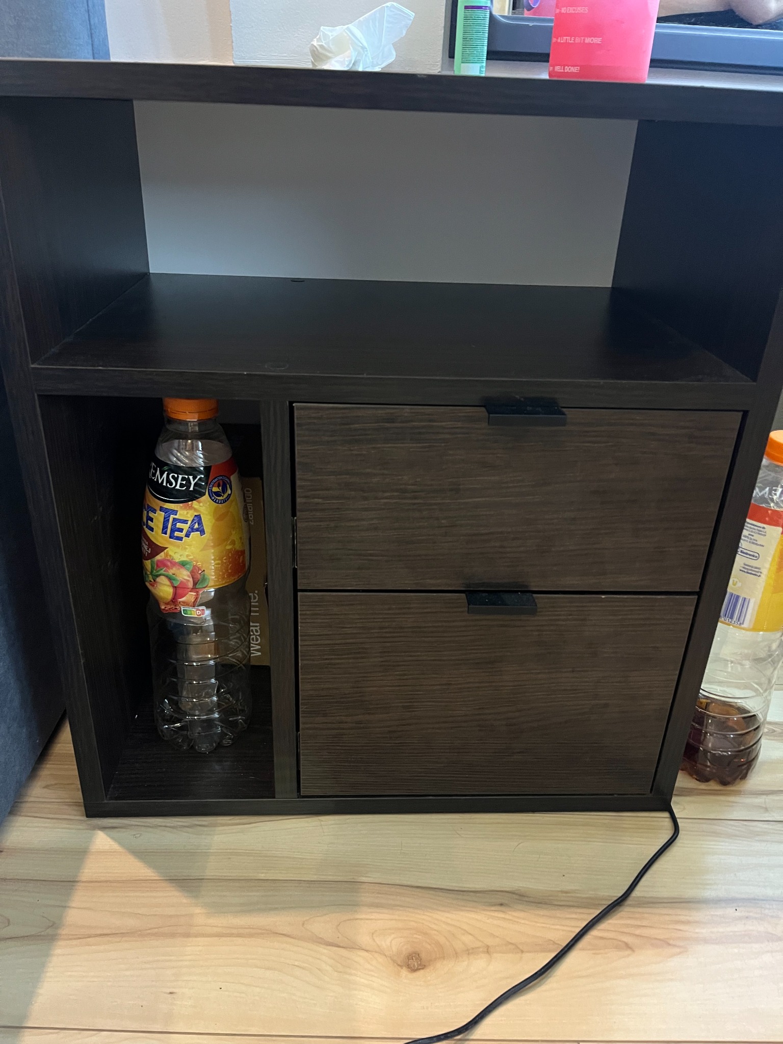 Ciemna szafka nocna z dwiema szufladami i otwartą półką, na której stoi butelka Ice Tea. Na blacie szafki widoczny różowy pojemnik.