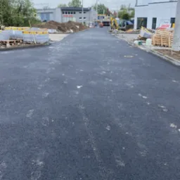 Nowa, asfaltowa droga dojazdowa z paletami kostki brukowej i sprzętem budowlanym w tle, przy pochmurnym niebie. Świeżo położony asfalt, widoczne ślady.