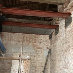 GDM CONSTRUCTION GRZEGORZ DZIADOWIEC - Ceglany budynek w trakcie rozbudowy. Widoczne stalowe belki, drewniane elementy konstrukcyjne i fragment rusztowania. Prace remontowe wewnątrz budynku.
