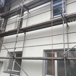 GDM CONSTRUCTION GRZEGORZ DZIADOWIEC - Świeżo otynkowana elewacja budynku w Brzesku, widoczna z poziomu rusztowania. Okna zabezpieczone folią. Jasna, gładka powierzchnia ściany.