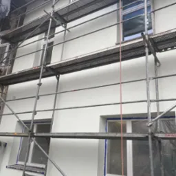 Świeżo otynkowana elewacja budynku w Brzesku, widoczna z poziomu rusztowania. Okna zabezpieczone folią. Jasna, gładka powierzchnia ściany.