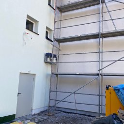 GDM CONSTRUCTION GRZEGORZ DZIADOWIEC - Elewacja budynku w trakcie prac z rusztowaniem; widoczne okna, drzwi i fragment instalacji. Na dole gruz i narzędzia budowlane.