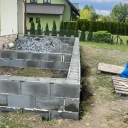 Budowa fundamentów z bloczków betonowych; widoczne szare bloczki, biała pianka montażowa, kamienie i zielony dom w tle.