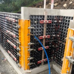 GDM CONSTRUCTION GRZEGORZ DZIADOWIEC - Szczegółowe ujęcie szalunku fundamentowego z żółtymi wzmocnieniami i niebieskim wężem, ukazujące etap przygotowania do wylania betonu na tle zieleni.