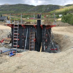 GDM CONSTRUCTION GRZEGORZ DZIADOWIEC - Szczegółowy widok szalunku zbrojeniowego w wykopie, przygotowanego do zalania betonem, z drabinkami i elementami konstrukcyjnymi na tle pagórkowatego krajobrazu.