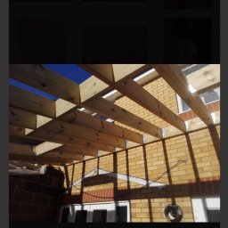 GDM CONSTRUCTION GRZEGORZ DZIADOWIEC - Drewniana konstrukcja z belek na tle ceglanego budynku i błękitnego nieba. Widoczne metalowe łączniki. Etap budowy zadaszenia tarasu lub werandy.