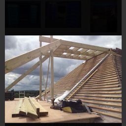 GDM CONSTRUCTION GRZEGORZ DZIADOWIEC - Konstrukcja drewniana dachu w trakcie budowy, widoczne krokwie, łaty, narzędzia i materiały budowlane. Tło stanowi krajobraz z zielenią i pochmurnym niebem.