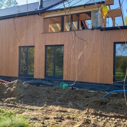 GDM CONSTRUCTION GRZEGORZ DZIADOWIEC - Nowoczesny dom z drewnianą elewacją i dużymi oknami, częściowo zasłonięty gałęziami drzew. Widoczne prace ziemne wokół budynku i instalacje.
