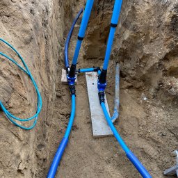 GDM CONSTRUCTION GRZEGORZ DZIADOWIEC - Niebieskie rury instalacji wodnej w wykopie ziemnym, połączone złączkami na drewnianych podkładkach. Widoczne ściany wykopu i fragment drabiny.