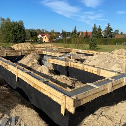 GDM CONSTRUCTION GRZEGORZ DZIADOWIEC - Zalane betonem szalunki fundamentów domu jednorodzinnego, zabezpieczone czarną folią. Widoczne zbrojenia i nasypany piasek. W tle zabudowa jednorodzinna i błękitne niebo.