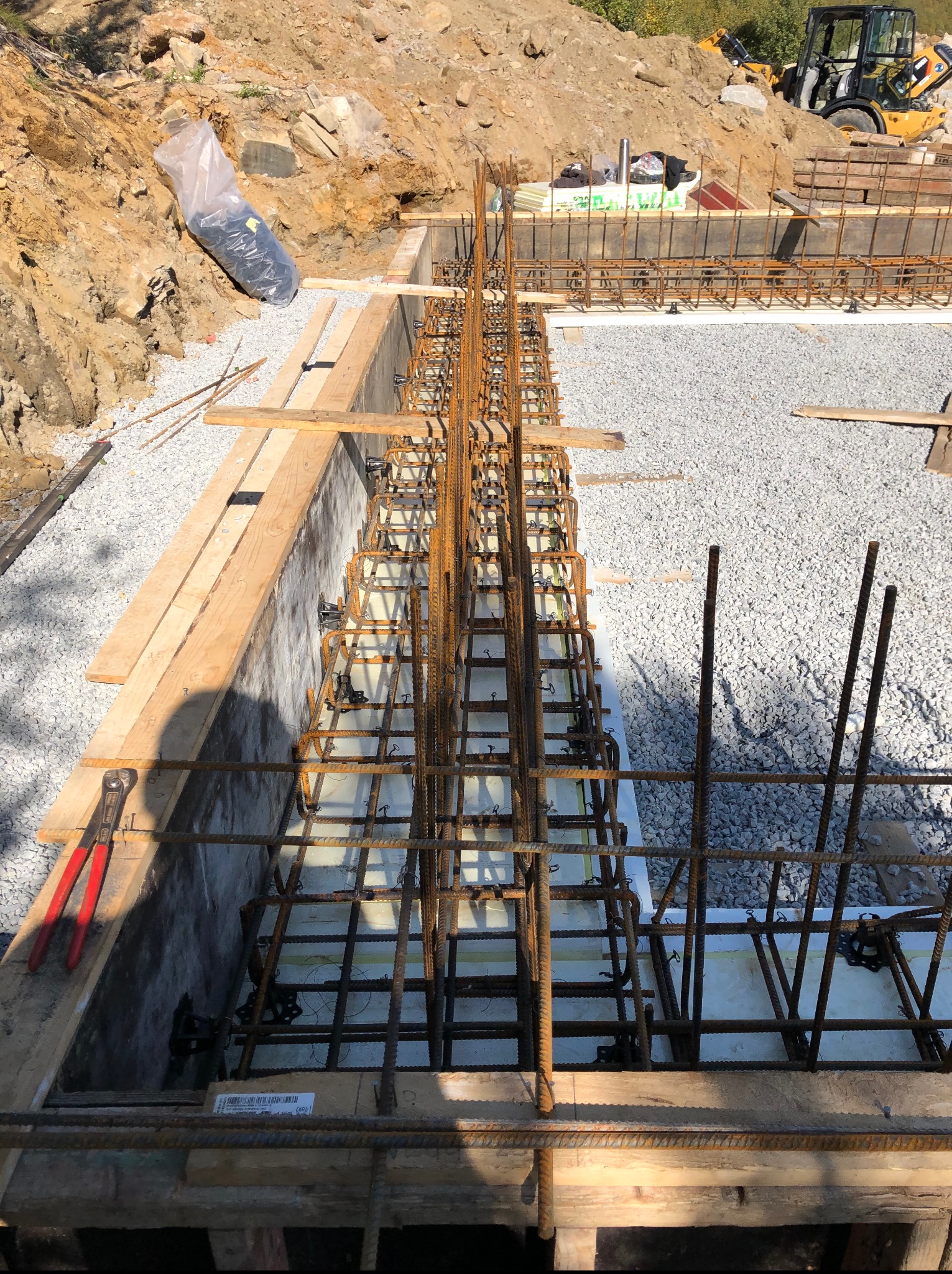 Zbrojenie fundamentów pod budowę domu, widok z góry. Konstrukcja z prętów stalowych w szalunku, przygotowana do zalania betonem. W tle wykop i maszyna budowlana.