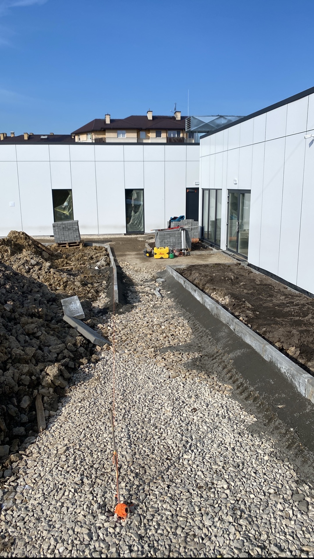 Przygotowanie terenu pod kostkę brukową przy nowoczesnym budynku z białymi panelami. Widoczne narzędzia i materiały budowlane.