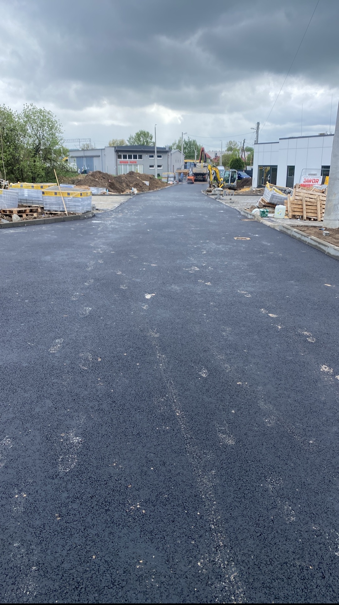 Nowa, asfaltowa droga dojazdowa z paletami kostki brukowej i sprzętem budowlanym w tle, przy pochmurnym niebie. Świeżo położony asfalt, widoczne ślady.