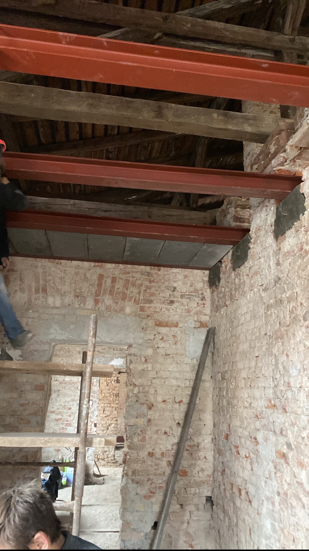 Ceglany budynek w trakcie rozbudowy. Widoczne stalowe belki, drewniane elementy konstrukcyjne i fragment rusztowania. Prace remontowe wewnątrz budynku.