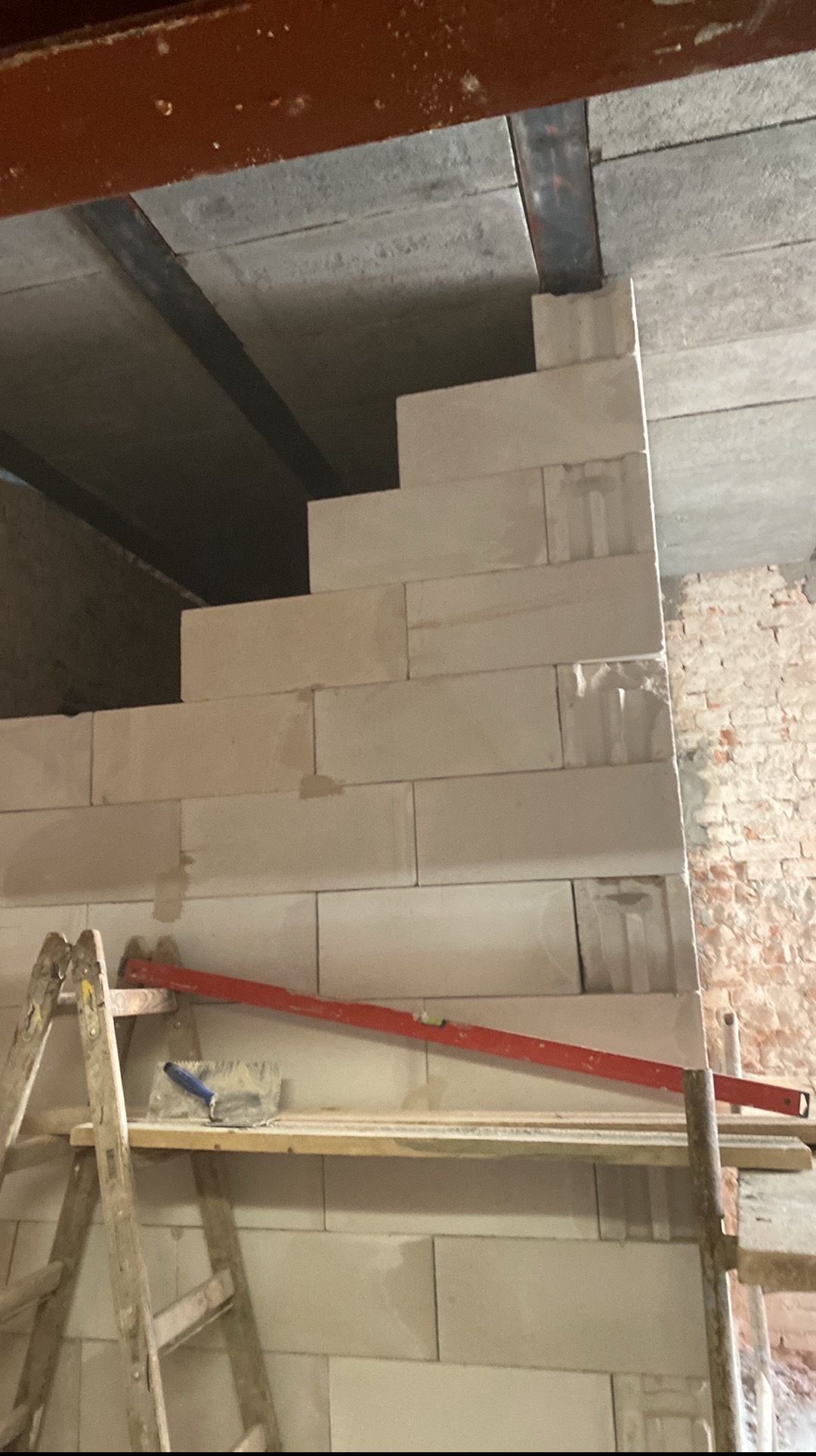 Wznoszenie ściany schodkowej z białych bloczków, widoczne rusztowanie, poziomica i kielnia. Fragment ceglanej ściany w tle. Budowa w trakcie realizacji.