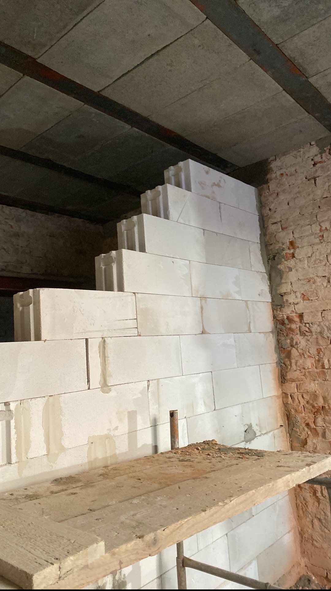 Wznoszenie ściany z bloczków betonu komórkowego na zaprawie, widoczne rusztowanie i fragment ceglanej ściany w tle. Budowa w trakcie realizacji.