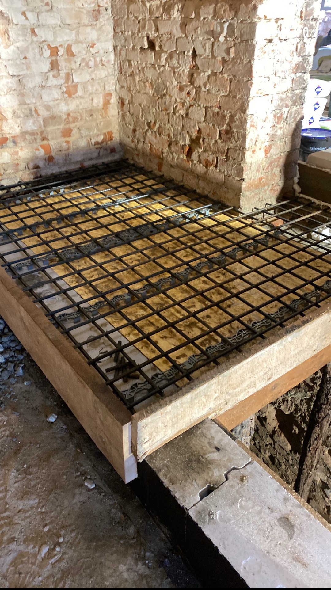 Zbrojenie pod wylewkę w drewnianej szalunku, widoczne zbrojenie siatkowe i dystanse, surowa ściana z cegły w tle. Etap prac przygotowawczych do wylewki.