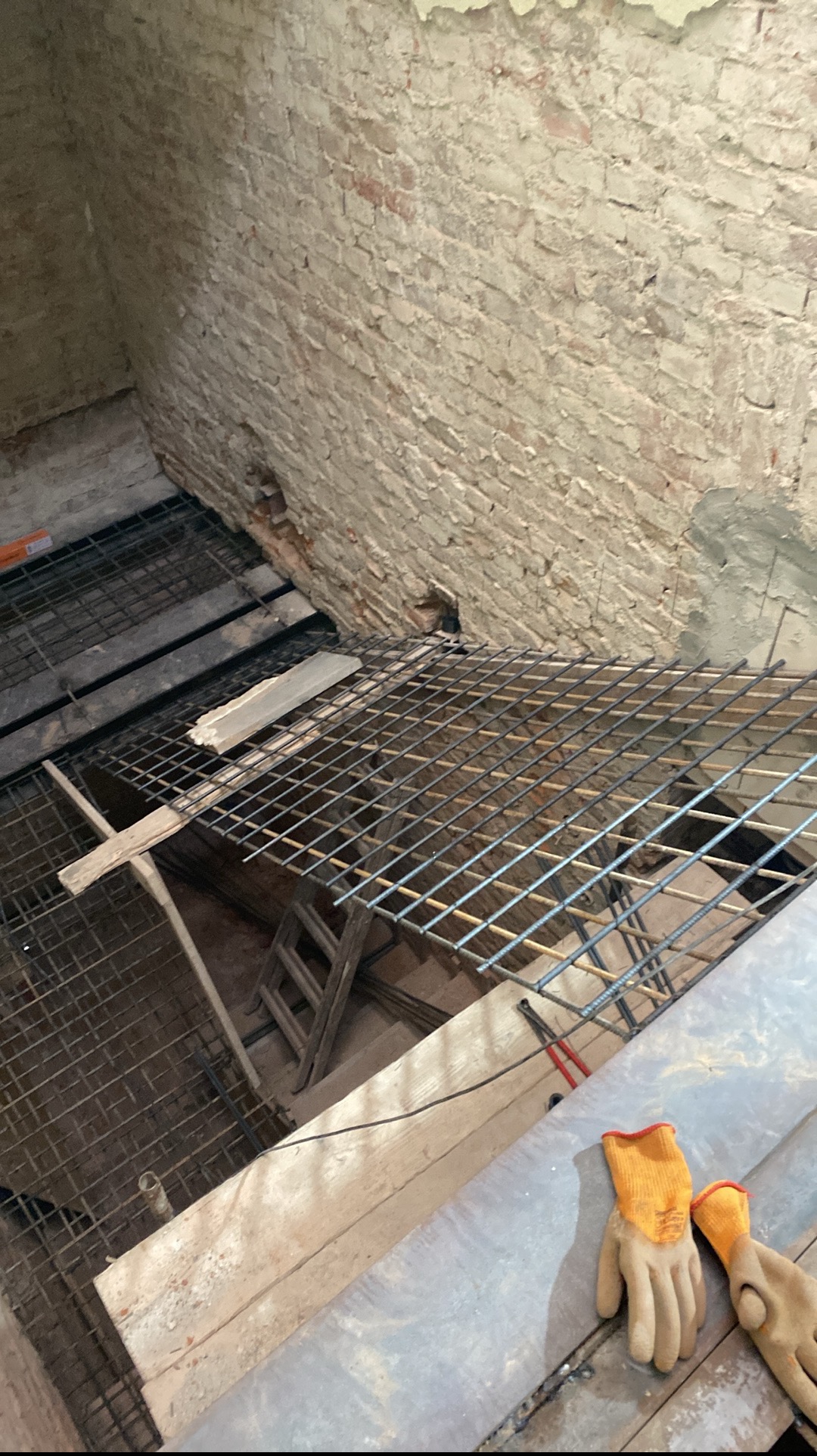Zbrojenie pod wylewkę w pomieszczeniu z ceglaną ścianą. Widoczne rękawice robocze, drewniane deski i fragment drabiny. Kąt ujęcia z góry.