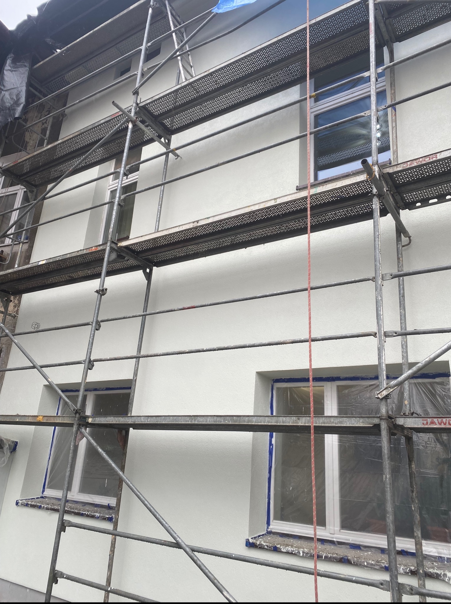 Świeżo otynkowana elewacja budynku w Brzesku, widoczna z poziomu rusztowania. Okna zabezpieczone folią. Jasna, gładka powierzchnia ściany.