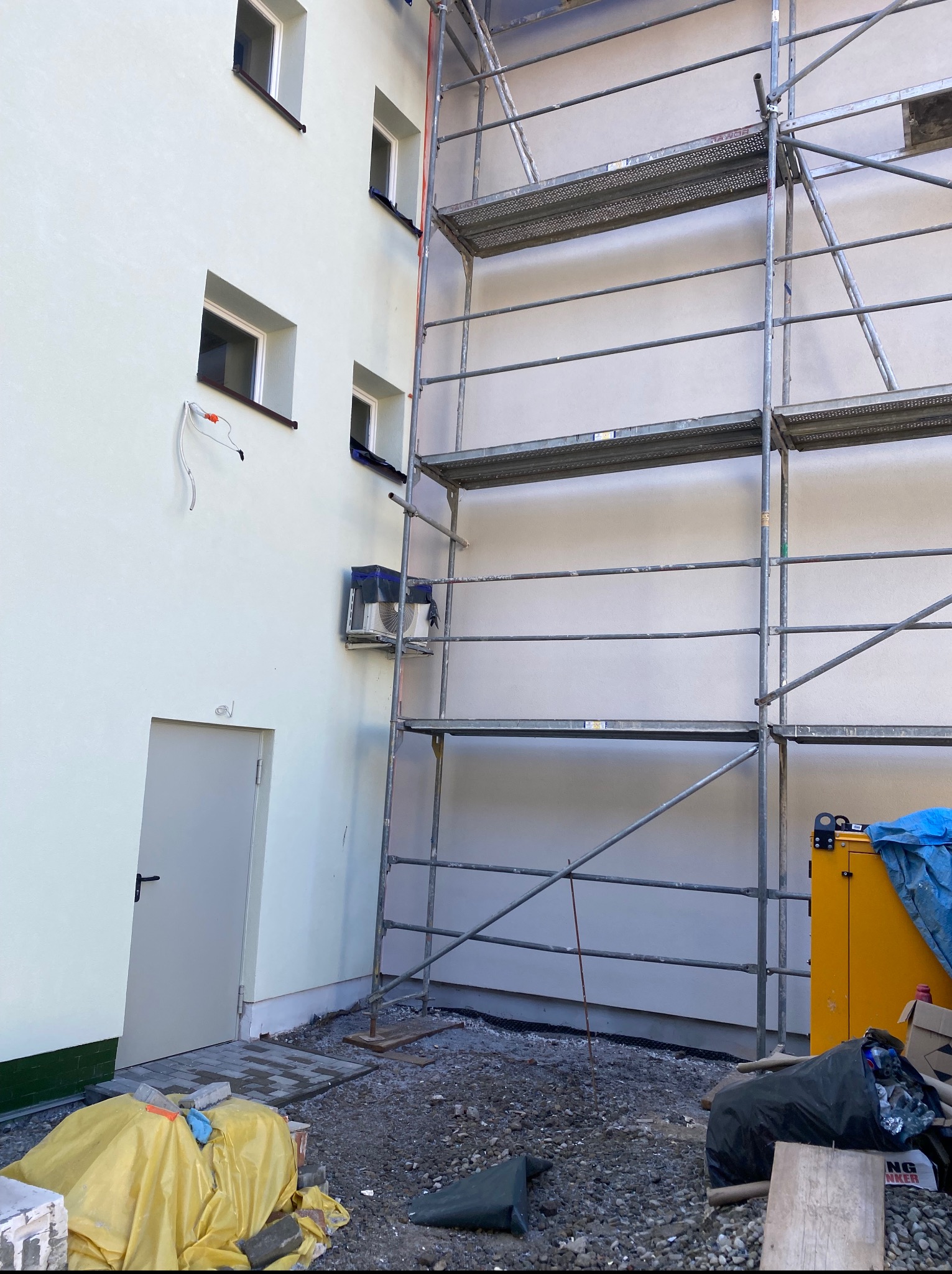 Elewacja budynku w trakcie prac z rusztowaniem; widoczne okna, drzwi i fragment instalacji. Na dole gruz i narzędzia budowlane.