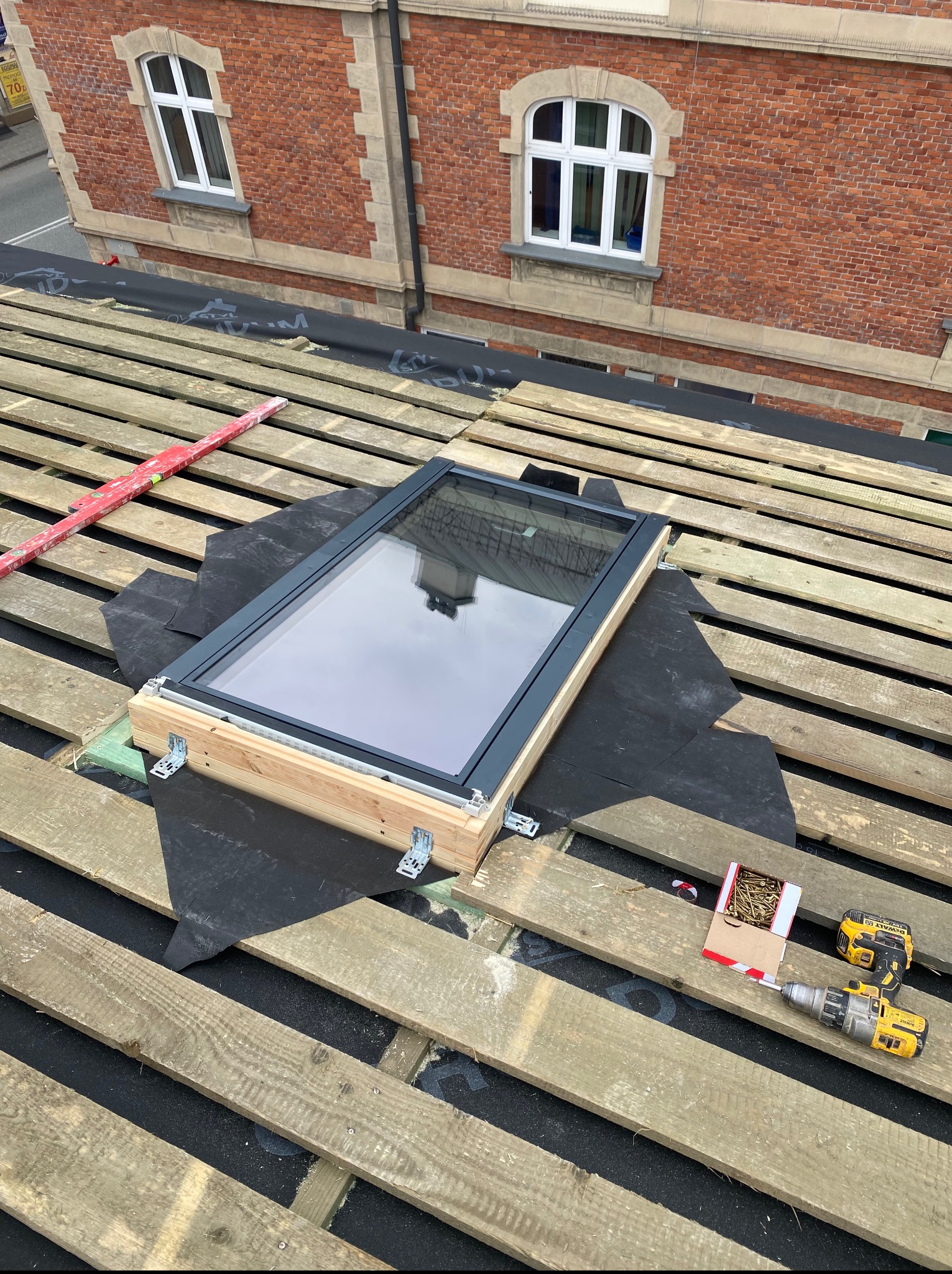 Montaż okna dachowego: drewniana konstrukcja, czarna membrana, narzędzia (wkrętarka, poziomica) na tle ceglanego budynku. Widoczny etap prac.